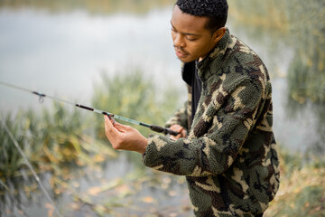 Hispanic man correcting fishing rod during fishing on river or lake coast at autumn season. Concept of rest, hobby and weekend in nature. Selective focus of male person in warm clothes