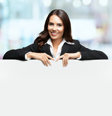 Happy smiling brunette business woman in confident black suit, standing behind, peeping from blank banner or mock up signboard, indoors. Posing in front of blurred modern office background.