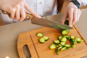 Close-up top view of unrecognizable woman cutting fresh cucumber cooking food salad sitting at wodden table in light kitchen room. Front view of female cooking vegetarian dieting salad at home.