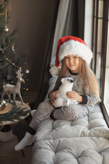 Full length cute little girl 3-4 year old in santa claus hat and pajamas sits at home on the windowsill by the window in beautiful grey interior decorated with a Christmas tree in background