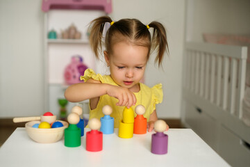 A two-year-old girl is playing in the children's room at a table with colorful cups and cylinders to study colors and logic. Children's educational toys