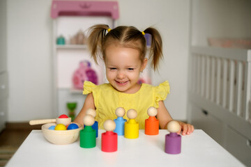 A two-year-old girl with ponytails is sitting at a table with developmental toys and laughing merrily. Bright colorful toys stand in front of the child. Baby is happy to learn colors