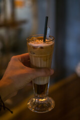 Close-up photo of glass cup with ice latte in a coffee shop. Hand holding the glass with iced caramel cappuccino with the straw in the restaurant.