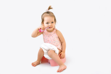 Сute girl is brushing teeth with a toothbrush while sitting on a potty. Baby does several things at the same time. Potty training concept. Toddler on a white studio background. Pink Children's potty