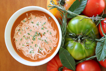 Bowl of tomato soup garnished with cream and chives surrounded by various tomatoes and herbs on wood background.