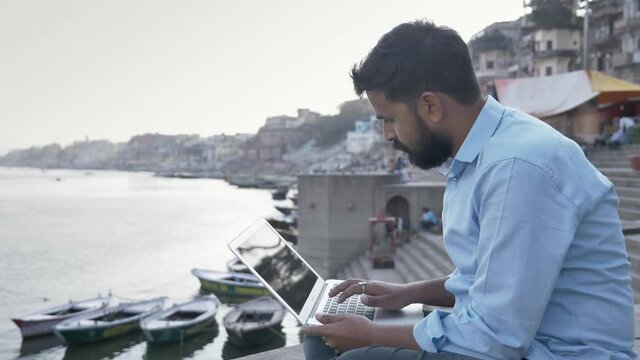 A movement shot of a young and handsome man is sitting on a platform working office projet using laptop next to the river, boats are seen in the background during sunset.