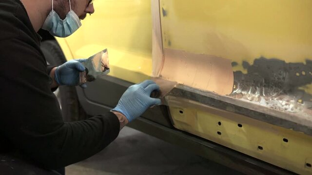 Auto mechanic repairing a yellow car with putty for paint in a workshop