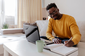 African young man in eyeglasses sitting at the table in front of laptop computer and looking at...