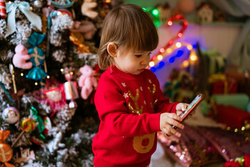 Fototapeta na wymiar Little girl in red sweater is watching cartoons on phone next to christmas tree at home while sitting on floor. Child waiting to receive present for christmas and new year. Kids and technology concept