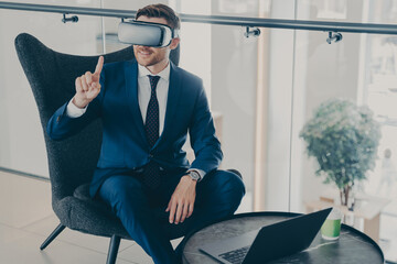 Smiling businessman using VR headset glasses while sitting in office lobby and working on laptop