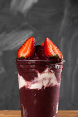 Brazilian Frozen Açai Berry Ice Cream Smoothie in plastic cup with Straw Berries and Condensed Milk. On a wooden desk and a gray summer background. Front view for menu and social media