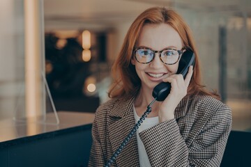 Female office worker in spectacles talking on landline phone with co-worker and smiling at camera