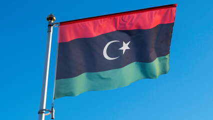 Naklejka na ściany i meble National flag of Islamic republic of Libya on a flagpole in front of blue sky with sun rays. Diplomacy concept.