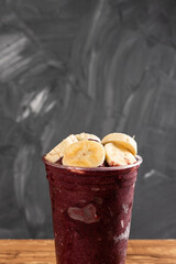 Brazilian Frozen Açai Berry Ice Cream Smoothie in plastic cup with Bananas. On a wooden desk and a gray summer background. Front view for menu and social media