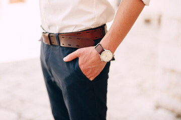 Man in a white shirt with sleeves rolled up, with a watch on his wrist, thrust his hands into the pockets of blue trousers with a brown belt. Close-up