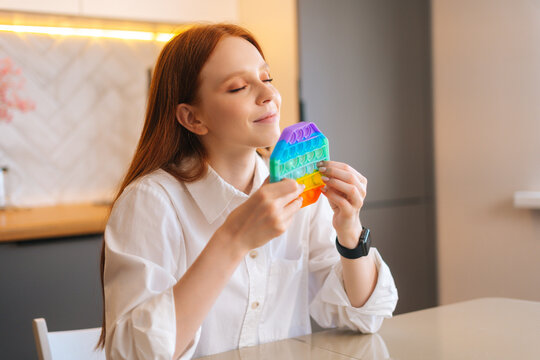 Close up face of happy charming redhead young woman pushing colorful iridescent soft silicone bubbles at home. Smiling female with trendy stress and anxiety relief fidgeting game in living room.