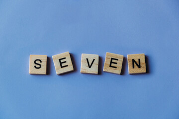 Number seven laid out from tiles in words. Wooden tiles on a blue background with letters. View from above. Copy space.