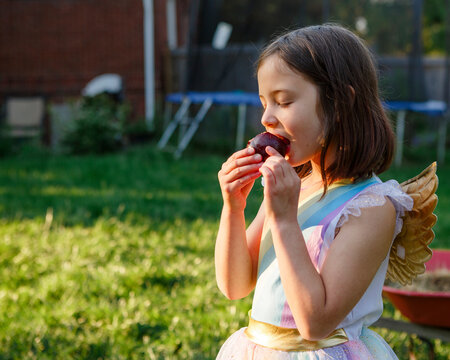 A small child in costume with golden wings eats juicy plum in sunshine