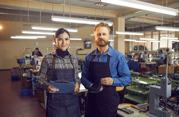 Portrait of two happy young shoe factory workers in aprons standing in big workshop, holding new...