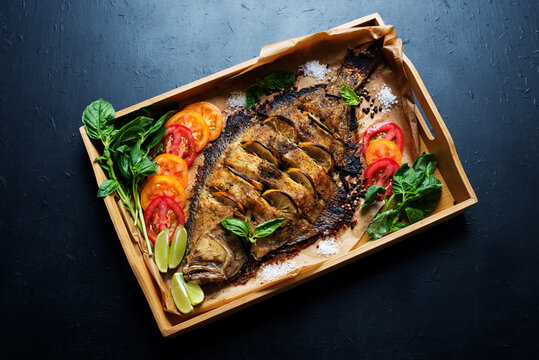 Oven-baked sea fish (flounder) on a bamboo tray on a dark background. Nearby are basil and tomatoes. Ready dish