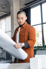 Fototapeta premium asian architect looking at blurred house models while holding blueprint in office