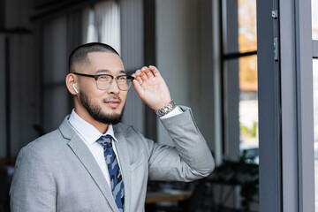positive asian businessman in earphone touching eyeglasses while looking away in office