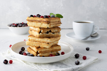 Viennese waffles with red and black currant berries lie on a white saucer. A light bowl with berries and coffee in a white cup in the background