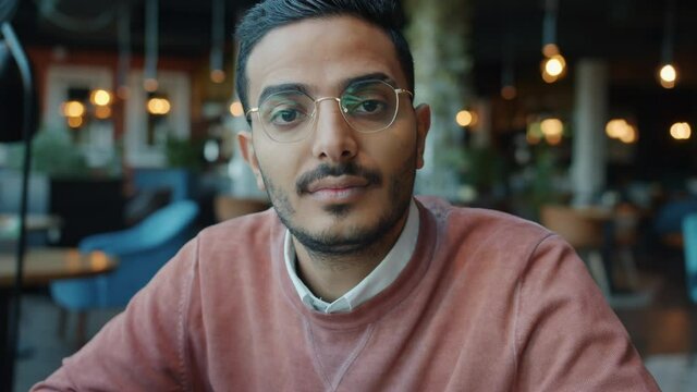 Slow motion portrait of serious Arab entrepreneur sitting in cafe alone wearing glasses and looking at camera with serious face. Youth and emotions concept.