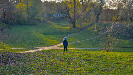 The boy stands at a fork in several paths that climb the hills. The rays of the setting sun lie on the grass and trees.