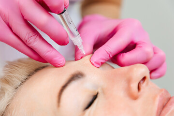 Obraz na płótnie Canvas Close-up portrait of beautiful adult woman getting injection in the cosmetology salon. Doctor in pink medical gloves with syringe injects filler in forehead