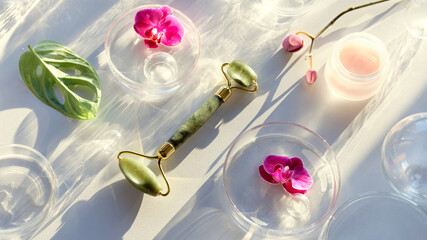 Jade stone face roller with monstera leaf, orchid flowers and glass baubles. Pink moisturizer cream in jar. Sunshine, long shadows. Beauty treatment flat lay on off white background with copy-space.