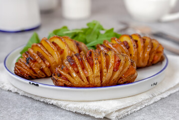 Bacon Hasselback Potatoes served on white plate with fresh arugula salad. Close up, selective focus, gray concrete background.