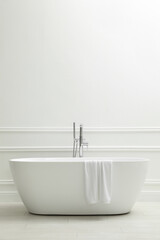 Modern ceramic bathtub with towel near white wall indoors, space for text