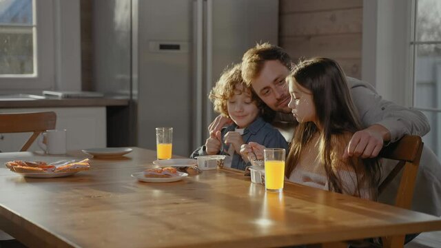 Man hugs his children drinking orange juice at breakfast on a sunny day. Happy family having fun at breakfast on a sunny day. 50fps. High quality 4k footage
