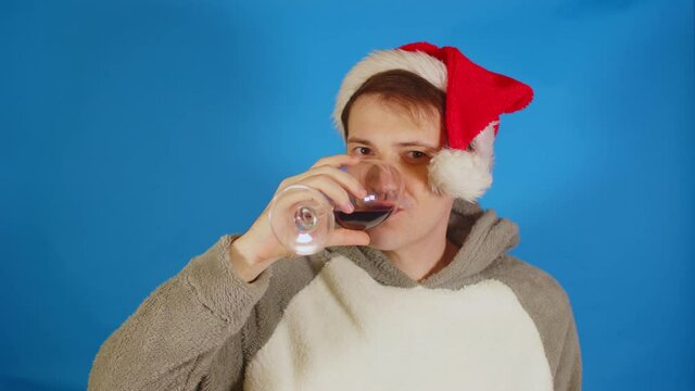 Happy man in santa hat with alcohol on blue background. Young male looking at camera, drinking red wine and smiling. Concept of celebrating holidays.