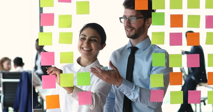 Multiethnic colleagues Indian and Caucasian teammates discuss collaborative project, jotting tasks writing ideas use colored sticky notes. Productive workflow, organizing thinking and planning concept