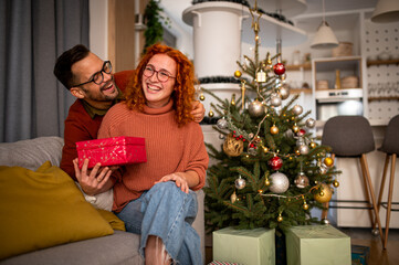 Obraz na płótnie Canvas Young couple in love sitting on a sofa next to a Christmas tree. The guy gives the woman a gift for the new year