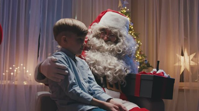 Little cute boy sitting near fireplace on Santa's knees, making a wish. Whitebearded Santa is hugging him and holding a gift.