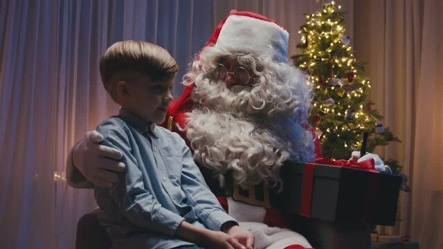 Little cute boy sitting near fireplace on Santa's knees, making a wish. Whitebearded Santa is hugging him and holding a gift.