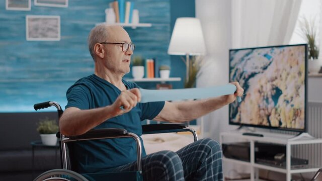 Elder person with disability stretching resistance band to exercise, looking at training lesson on tablet. Senior man sitting in wheelchair and pulling elastic belt to train with workout video