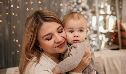 tender photo of a blonde mom holding a baby baby against the background of a Christmas tree
