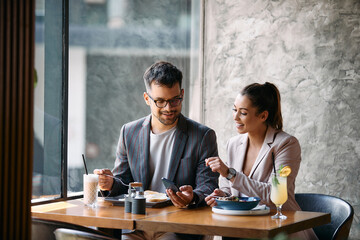 Young happy colleagues use mobile phone while having breakfast in cafe.