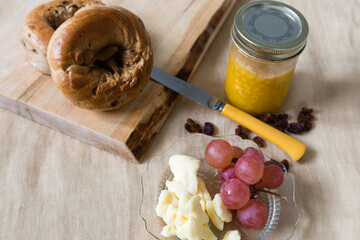 High angle selective focus view of healthy foods, including bagels, homemade jam, grapes, cheese curds and raisins on linen tablecloth