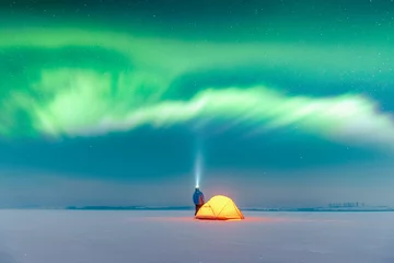  Tourist with flashlight near yellow tent lighted from the inside against the backdrop of incredible starry sky with Aurora borealis. Amazing night landscape. Northern lights in winter field © Ivan Kmit