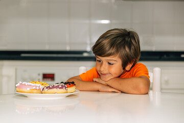 Blond boy sitting in the kitchen in front of four colorful donuts