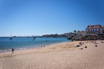 Fototapeta na wymiar Beautiful Cascais Beach with people in the sand and beach houses and white boats in the distance over the beautiful blue sky and blue sea on a sunny spring day. 