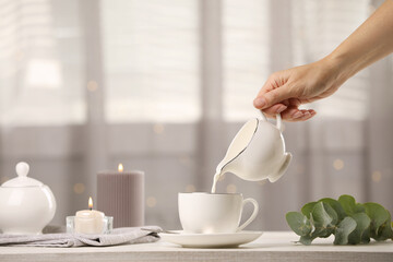 Woman pouring milk into cup at table indoors, closeup