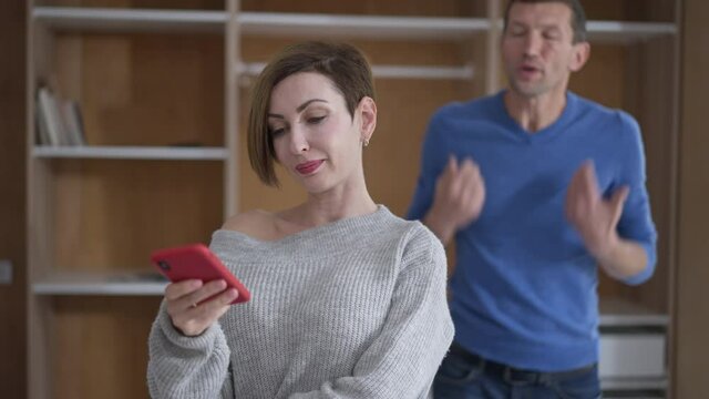 Portrait of egoistic wife with vainglorious facial expression surfing social media on smartphone as blurred husband screaming gesturing at background. Disinterested Caucasian woman ignoring man