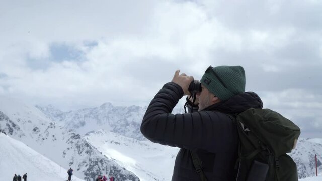 Man in warm jacket and with backpack looking away with binocular in the snowy mountains