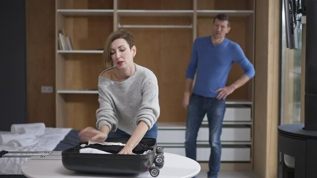 Portrait of stressed Caucasian attractive woman packing travel bag with blurred shocked man standing at background in bedroom talking. Unhappy wife leaving husband. Divorce concept